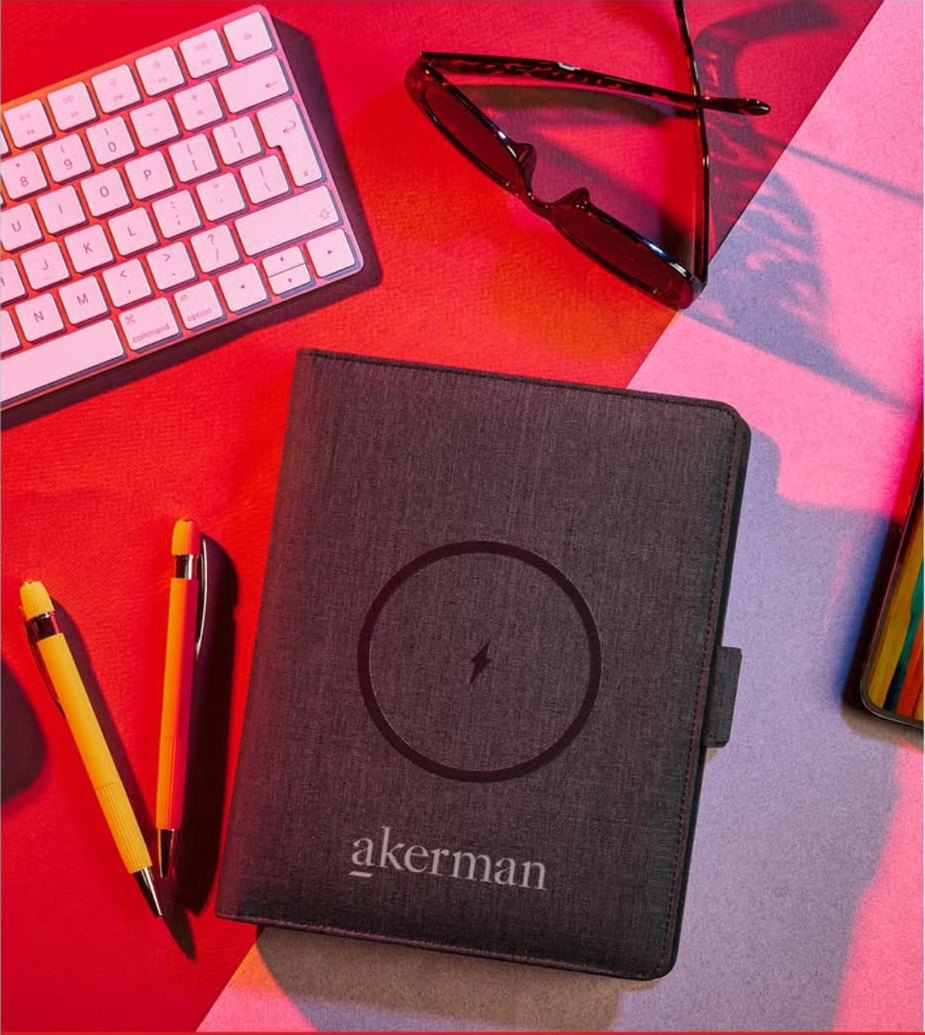 Dark fabric wireless-charging portfolio with a company logo, beside pens and a keyboard